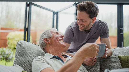 two people interacting in living room one sitting on couch holding mug other standing leaning towards seated individual